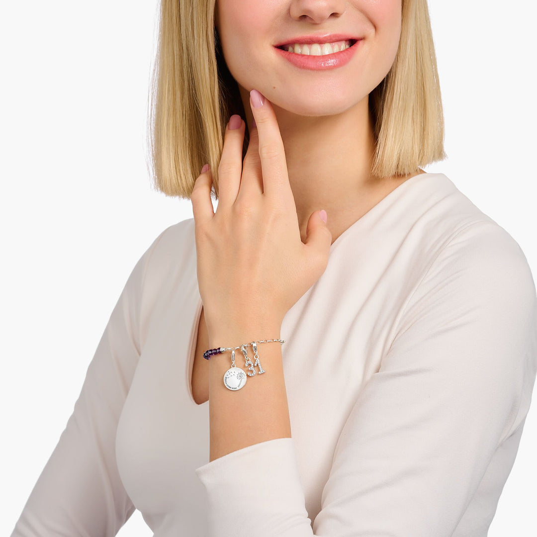 Woman wearing a THOMAS SABO sterling silver charm bracelet with purple beads, a dandelion disc charm, and crystal number charms.
