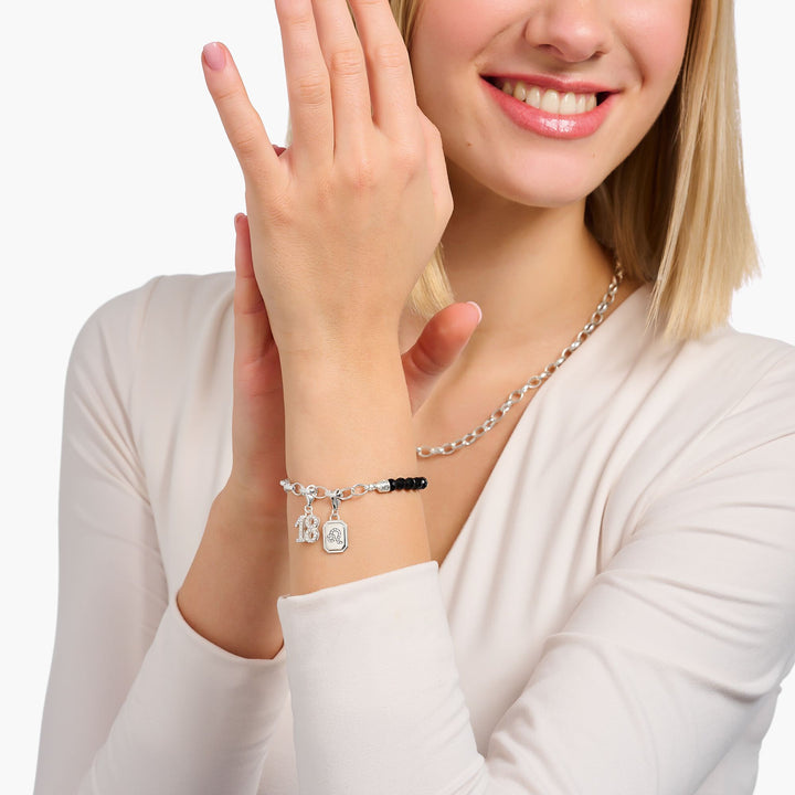 Woman wearing a sterling silver charm bracelet with black beads, "18" charm and square pendant, paired with chunky chain necklace.