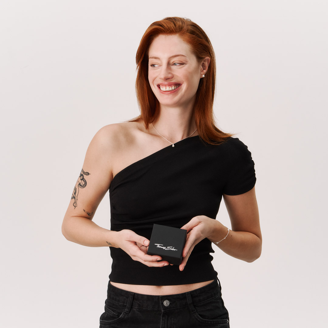 Smiling woman holding a THOMAS SABO gift box, wearing a delicate silver necklace with pearl pendant and silver bracelet.