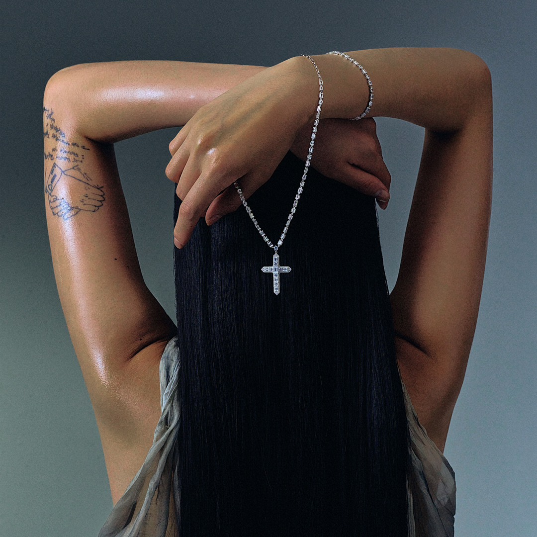 Woman seen from behind showcasing a THOMAS SABO crystal-pavé cross pendant necklace and silver bracelets, with dark hair and arm tattoo.