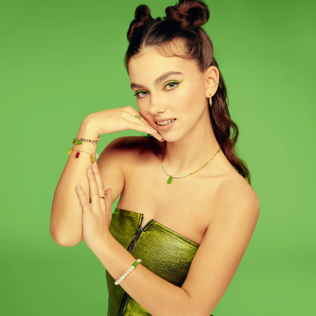 Hand holding a green apple, styled with THOMAS SABO gummy bear charms, pearl, crystal tennis, and rainbow stone gold-plated bracelets and rings.
