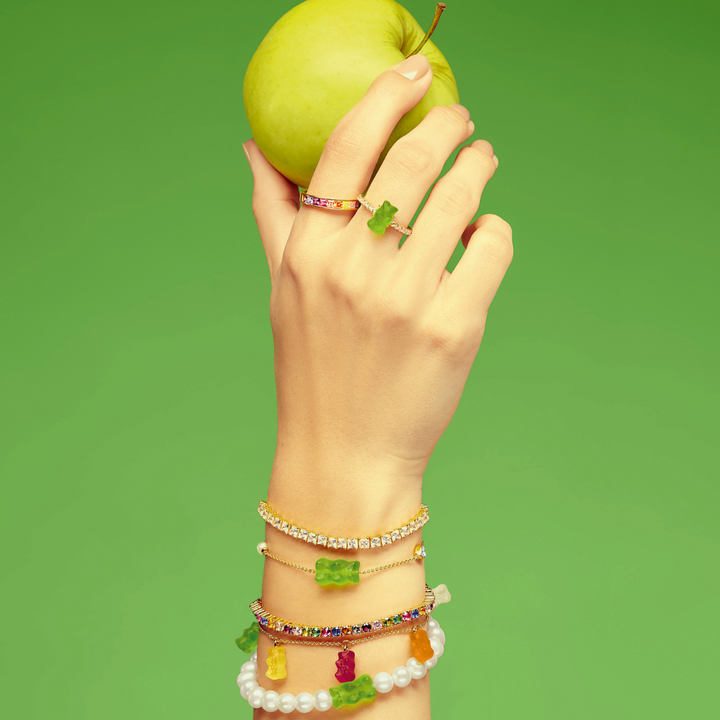 Hand holding a green apple, styled with THOMAS SABO gummy bear charms, pearl, crystal tennis, and rainbow stone gold-plated bracelets and rings.