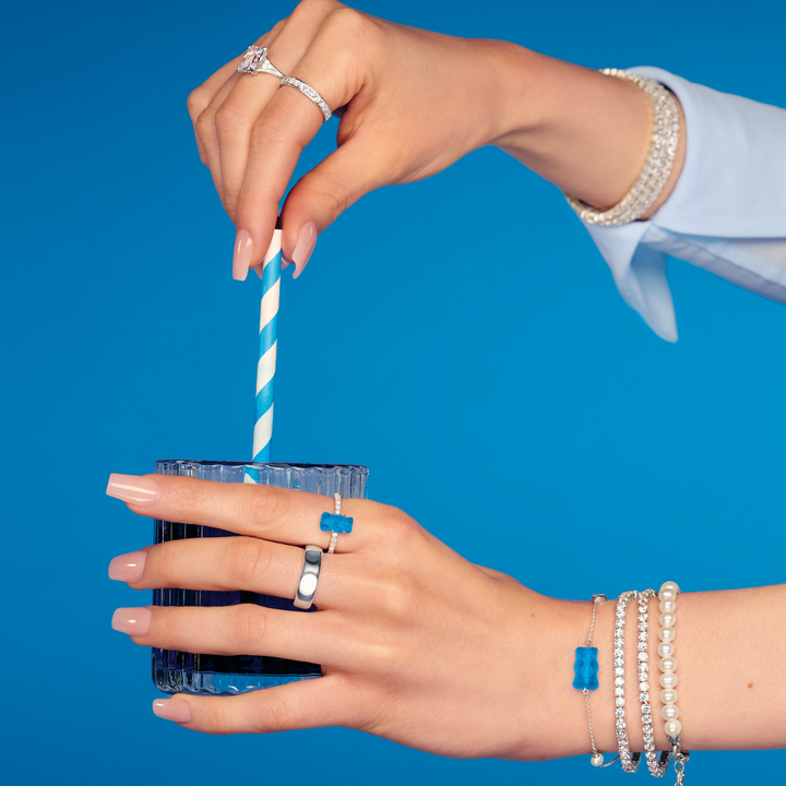 Hand holding a green apple, styled with THOMAS SABO gummy bear charms, pearl, crystal tennis, and rainbow stone gold-plated bracelets and rings.