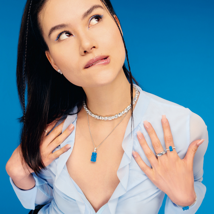 Hand holding a green apple, styled with THOMAS SABO gummy bear charms, pearl, crystal tennis, and rainbow stone gold-plated bracelets and rings.