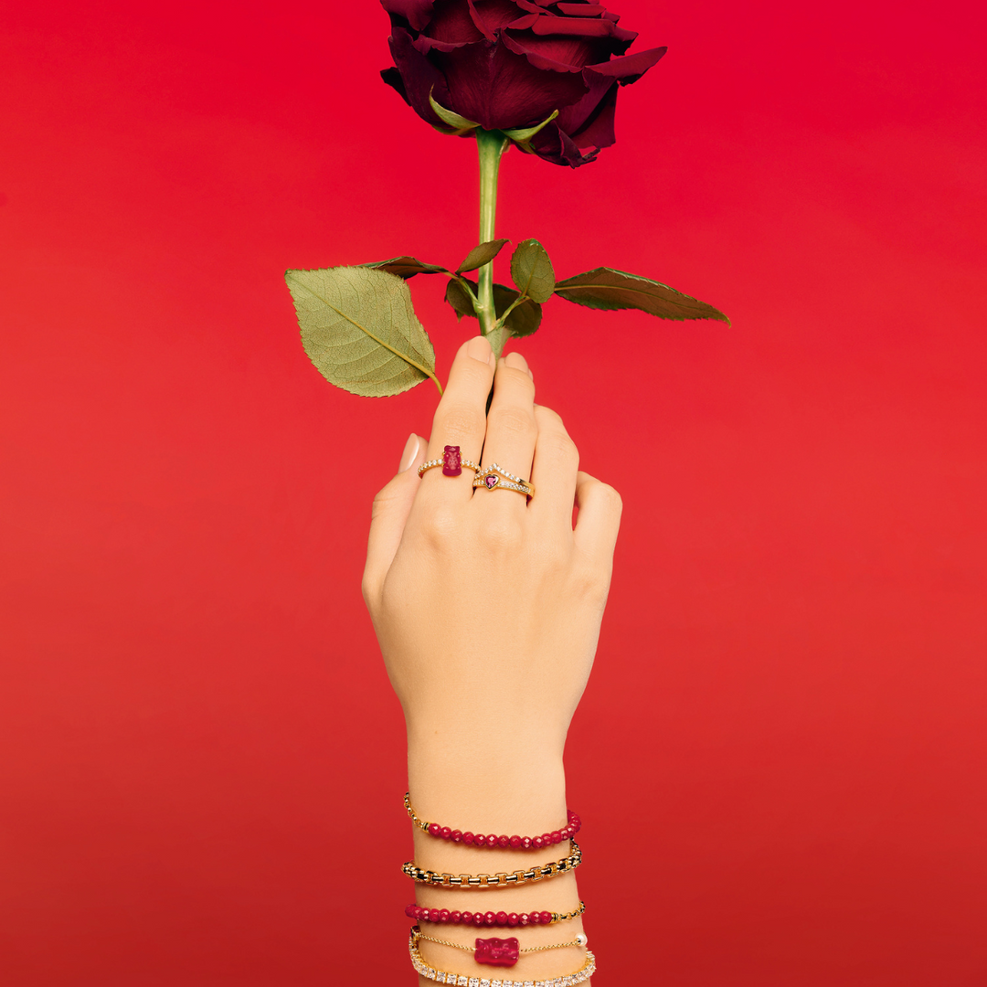 Hand holding a green apple, styled with THOMAS SABO gummy bear charms, pearl, crystal tennis, and rainbow stone gold-plated bracelets and rings.