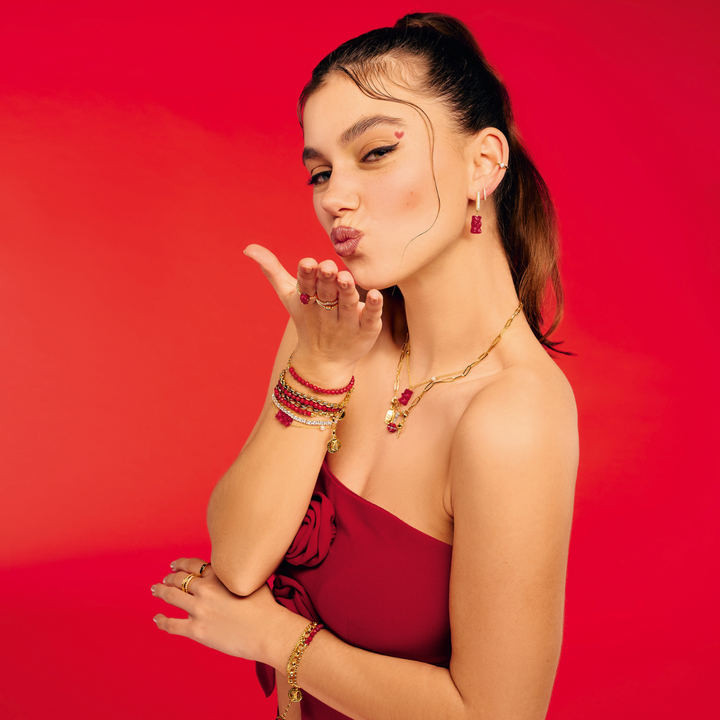 Hand holding a green apple, styled with THOMAS SABO gummy bear charms, pearl, crystal tennis, and rainbow stone gold-plated bracelets and rings.
